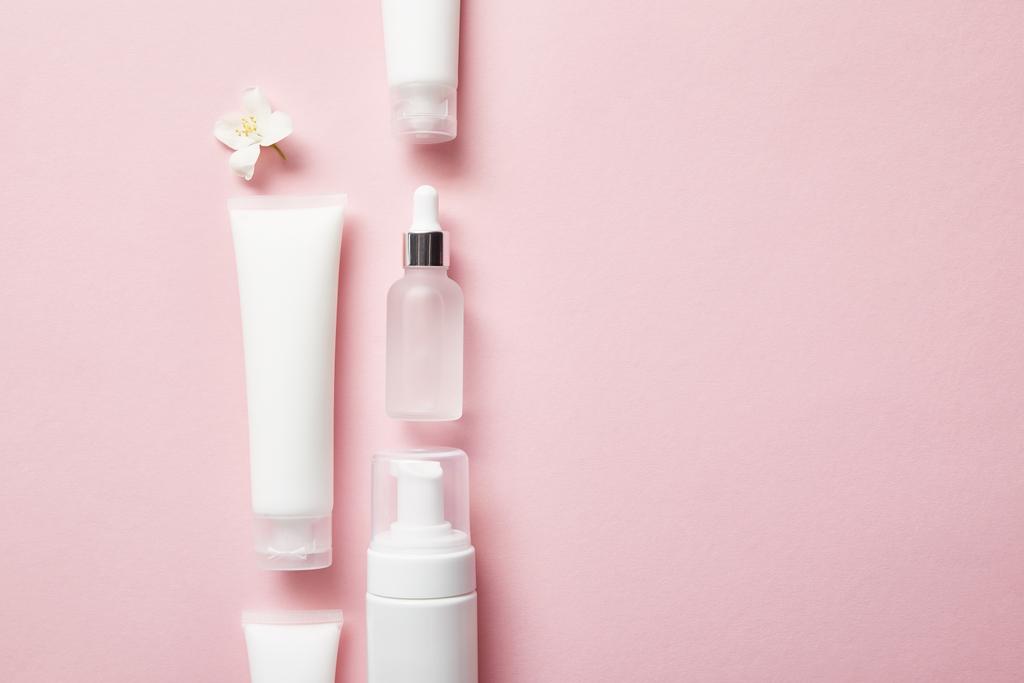 platte lay van cosmetische glazen fles, crème buizen met room, cosmetische dispenser en jasmijn bloem op roze  - Foto, afbeelding
