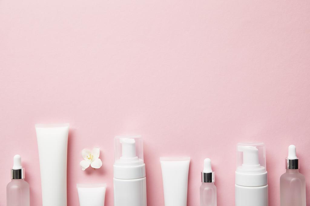 Draufsicht auf kosmetische Glasflaschen, Cremetuben mit Creme, Kosmetikspender und Jasminblüte auf rosa  - Foto, Bild