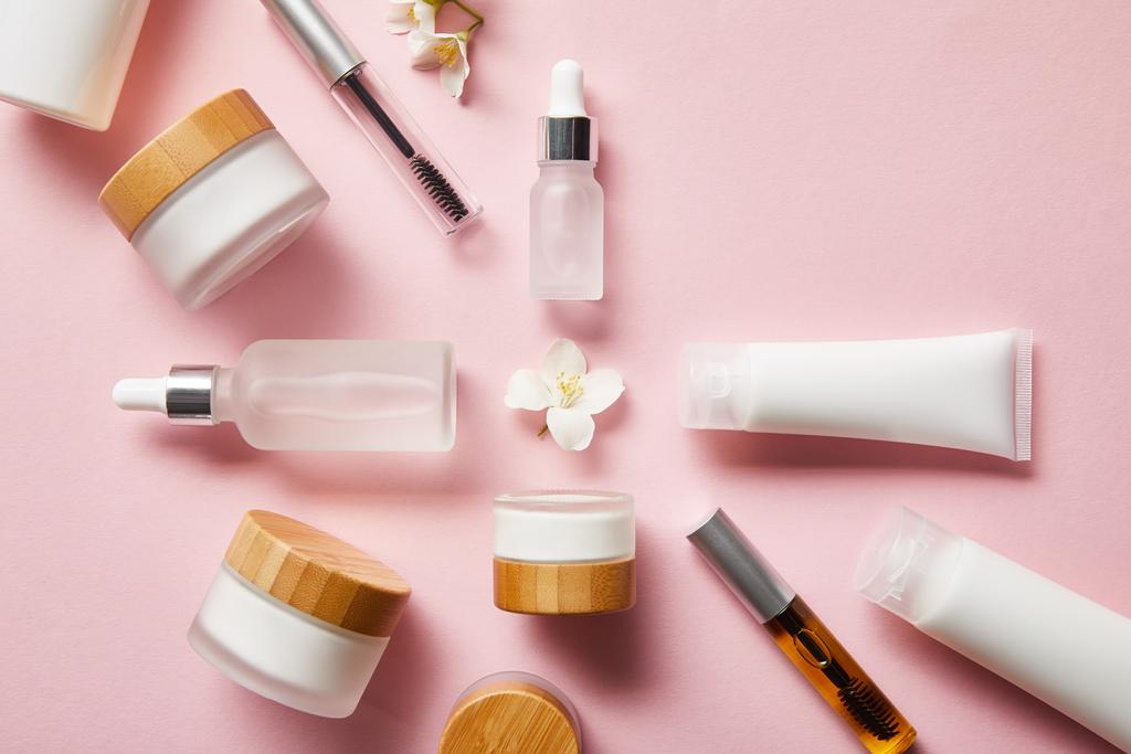 Draufsicht auf Gläser, Cremetuben mit Handcreme, Kosmetikglasflaschen, Mascaraflaschen und Jasminblüte in der Mitte auf rosa  - Foto, Bild