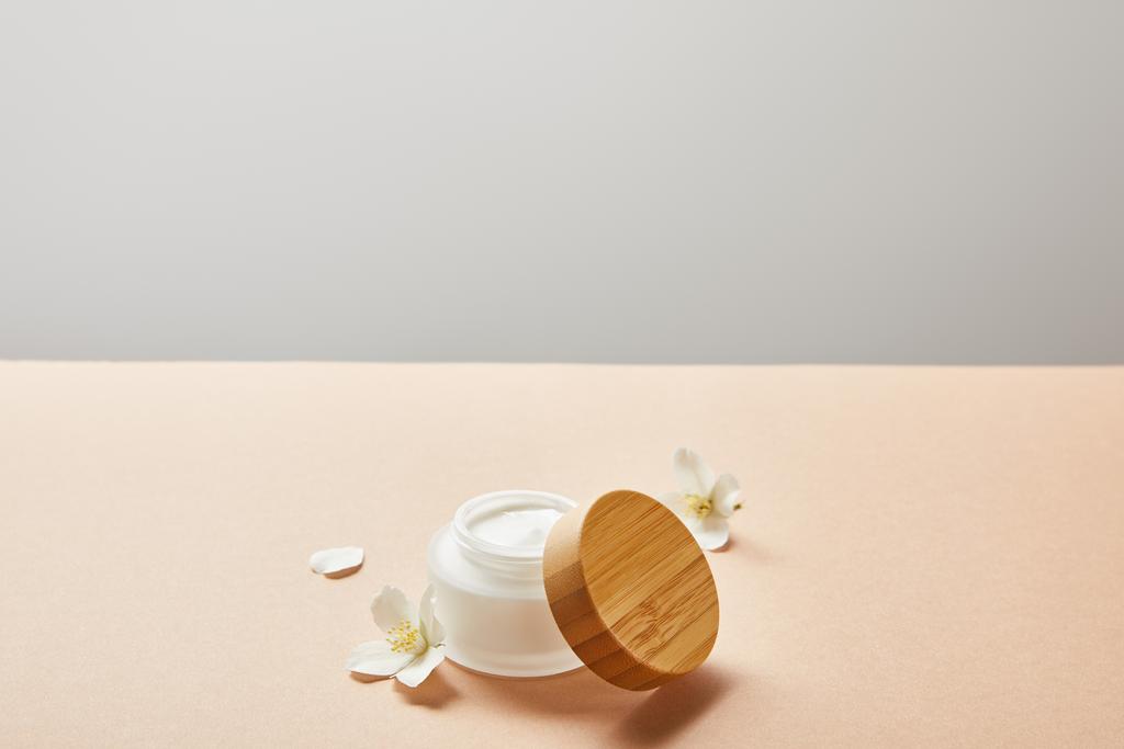 open pot met gezichtscrème en enkele jasmijn bloemen op beige geïsoleerd op grijs - Foto, afbeelding