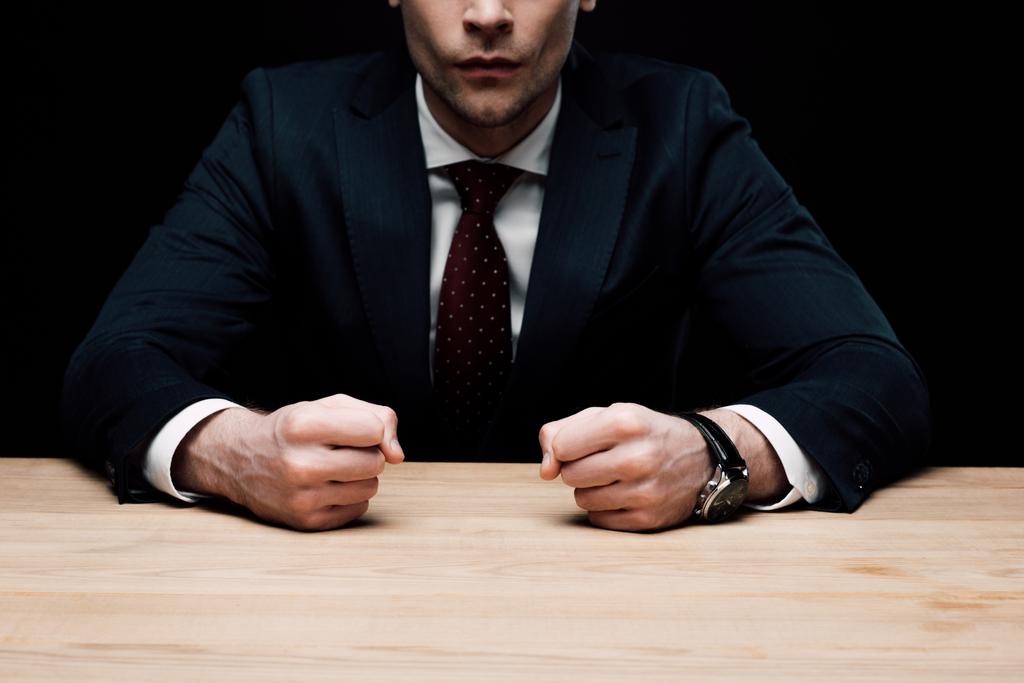 partial view of angry businessman sitting at table and holding fists isolated on black, human emotion and expression concept - Photo, Image