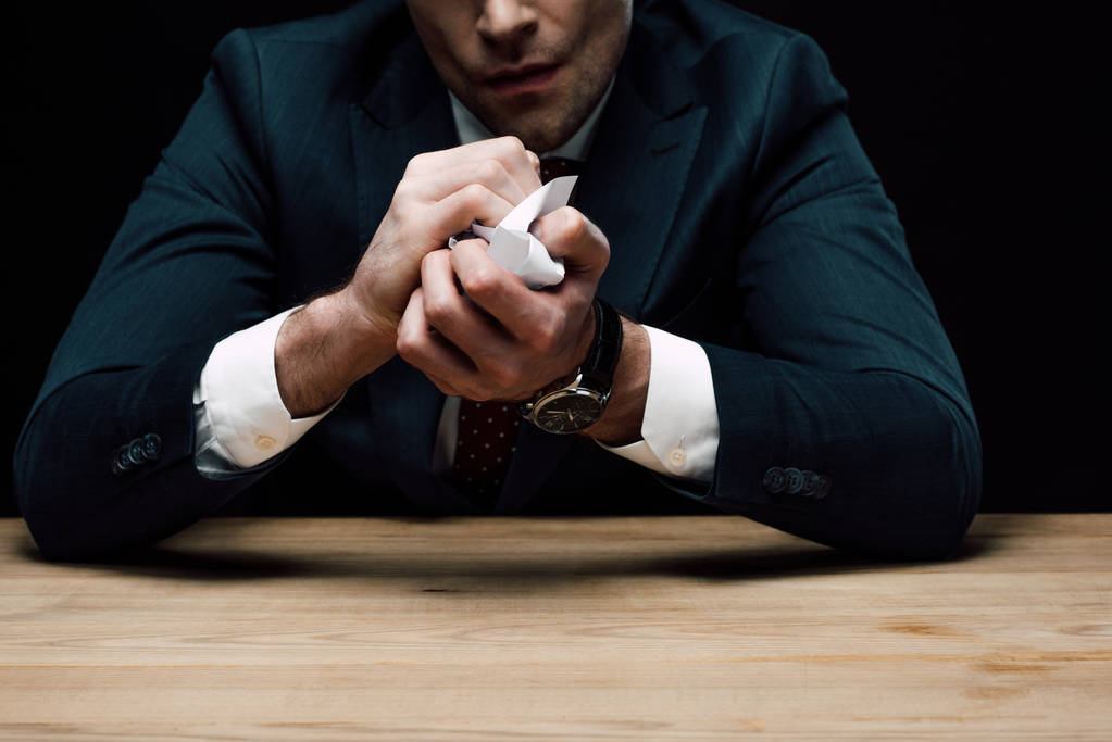 Teilansicht eines depressiven Geschäftsmannes, der Papier zerknüllt, während er isoliert auf schwarzem Papier am Holztisch sitzt - Foto, Bild
