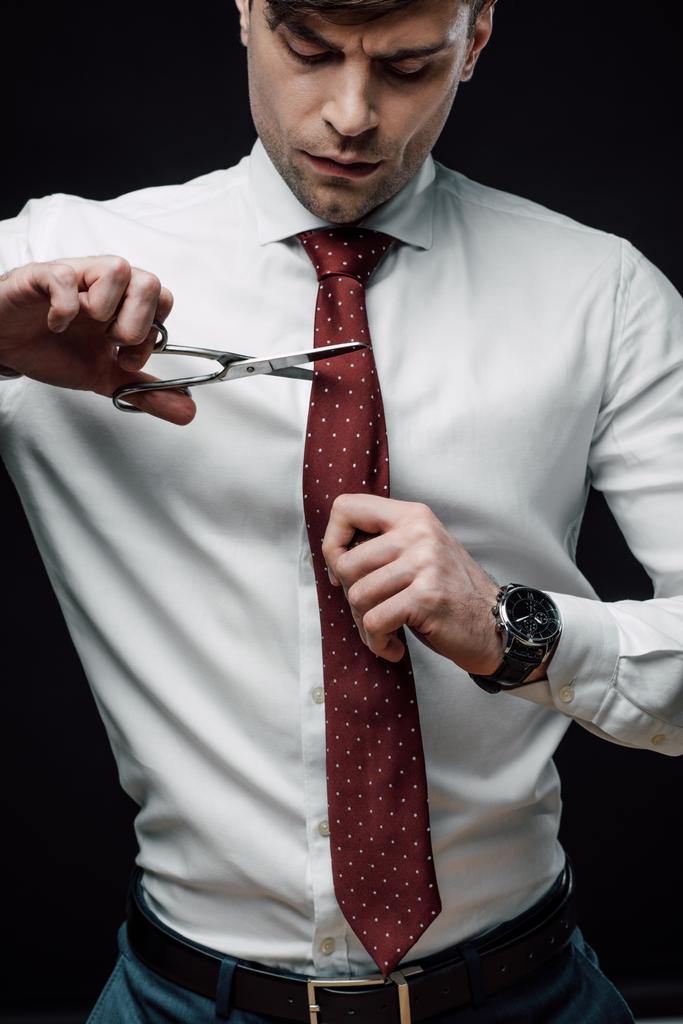 αναστατωμένος επιχειρηματίας κοπή γραβάτα με ψαλίδι απομονώνεται σε μαύρο - Φωτογραφία, εικόνα