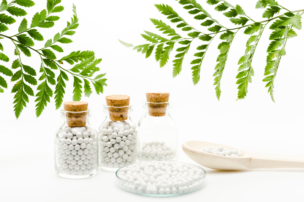 selective focus of small pills in glass bottles with wooden corks near spoon and green leaves on white  - Photo, Image