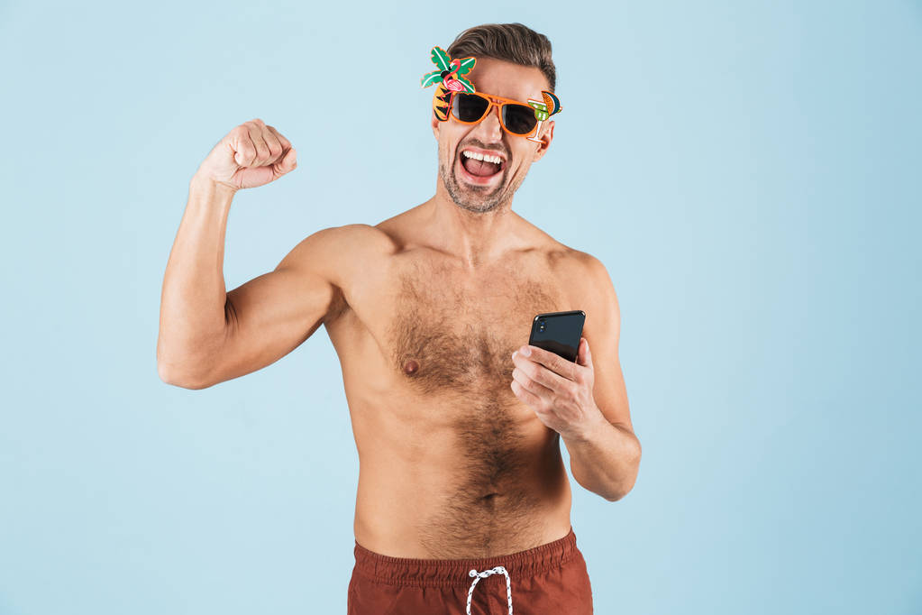 Возбужденный счастливый взрослый мужчина в купальниках позирует изолированно на фоне синей стены с помощью мобильного телефона
. - Фото, изображение