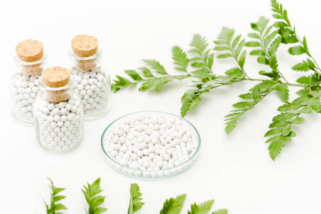 επιλεκτική εστίαση των στρογγυλά χάπια σε γυάλινη πλάκα κοντά σε μπουκάλια με ξύλινα φελλούς και πράσινα φύλλα σε λευκό  - Φωτογραφία, εικόνα