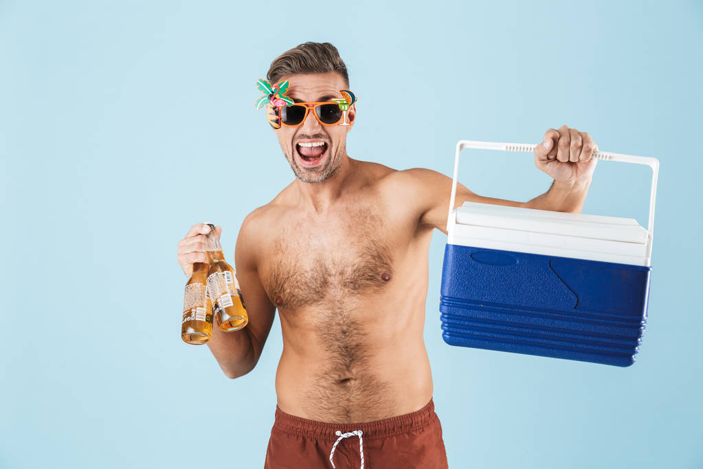 Захоплений щасливий дорослий чоловік у купальнику позує ізольовано на синьому фоні стіни, тримаючи холодильник з пивом у сонцезахисних окулярах
. - Фото, зображення