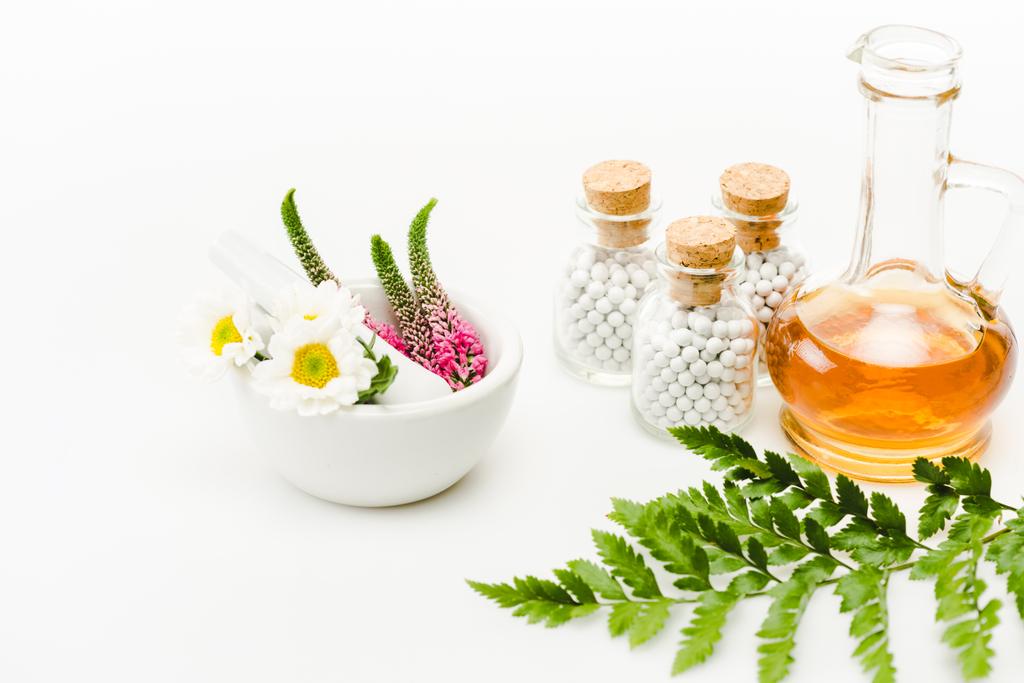 bloemen in mortel in de buurt van stamper en glazen flessen met pillen, kruik met olie en groen blad op wit  - Foto, afbeelding