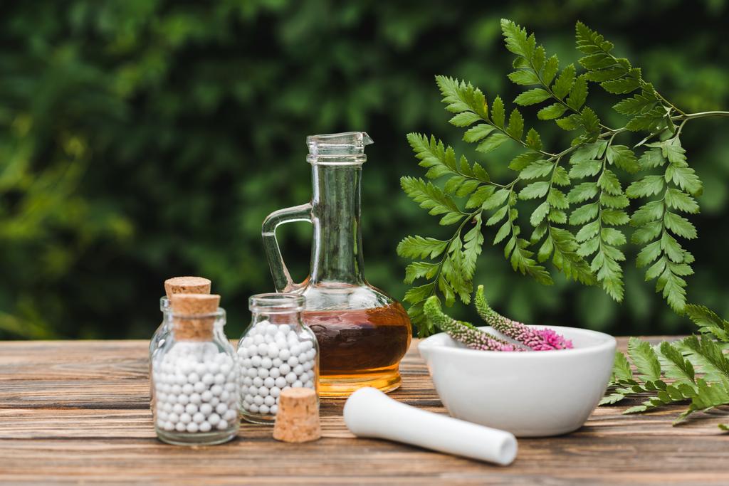 selective focus of pestle near mortar with flowers, glass bottles with pills and jug with oil on wooden table  - Photo, Image