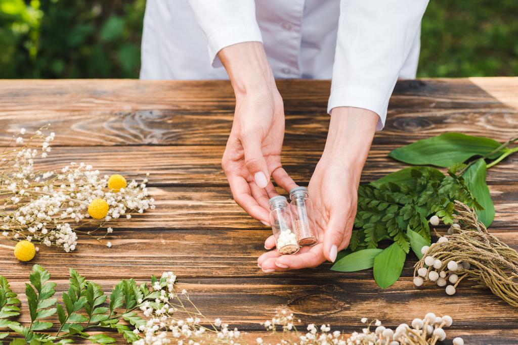 vista cortada da mulher segurando garrafas de vidro com pílulas perto de plantas
  - Foto, Imagem