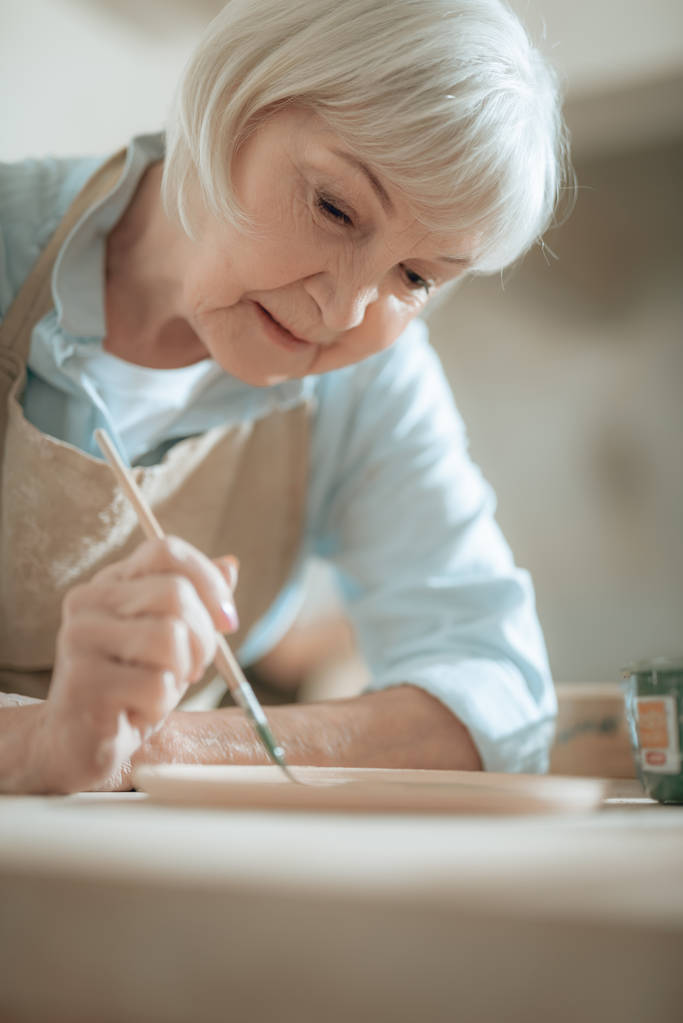 Обрезанное фото пожилой ремесленницы, рисующей декоративную тарелку в мастерской
 - Фото, изображение