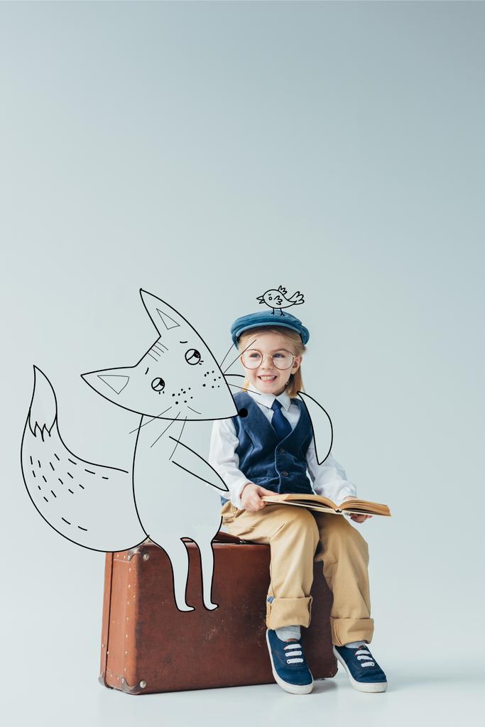 χαμογελαστή παιδί σε ρετρό γιλέκο με φαντασία πουλί στο καπάκι κάθεται σε βαλίτσα κοντά νεράιδα αλεπού και κρατώντας το βιβλίο σε γκρίζο φόντο - Φωτογραφία, εικόνα