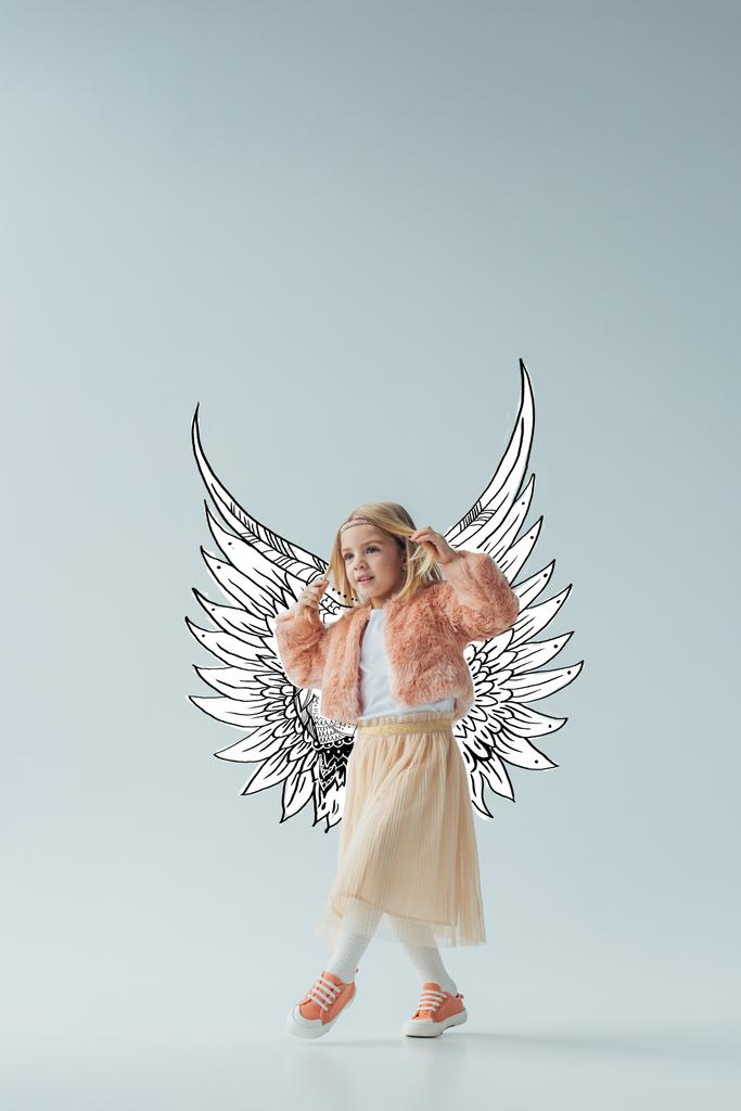 lindo niño en abrigo de piel sintética y falda con alas de ángel de hadas caminando y mirando hacia otro lado sobre fondo gris
 - Foto, imagen