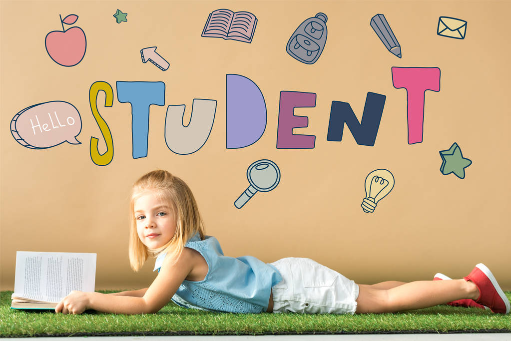 cute kid lying on grass rug and holding book on beige background with student lettering - Photo, Image