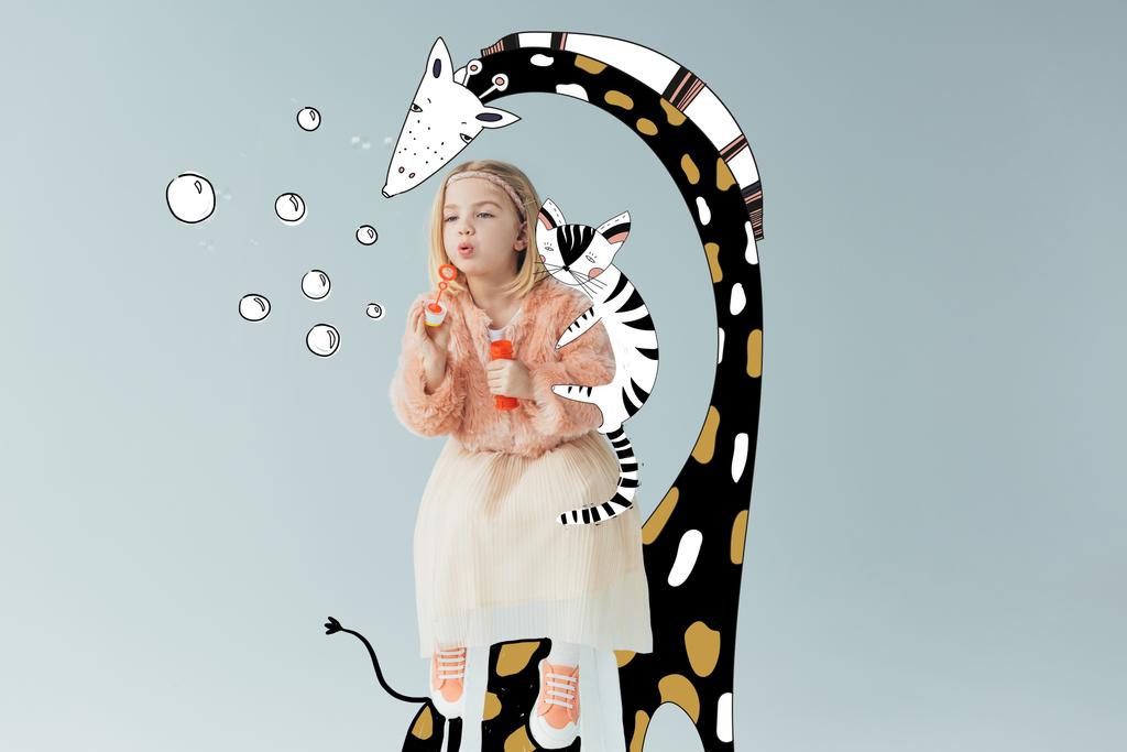 schattig kind in Faux Fur jas en rok zittend op Fairy Giraffe en blazen zeepbellen geïsoleerd op grijs - Foto, afbeelding