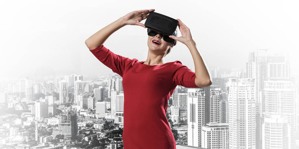 Horizontální záběr mladé a krásné ženy v červených šatech s použitím sluchátka s virtuální realitou, která stojí venku s moderním výhledem na pozadí - Fotografie, Obrázek