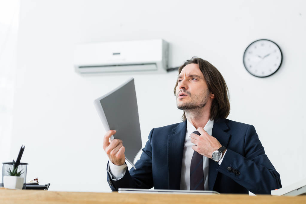 hombre de negocios sentado en la oficina con la carpeta en la mano, mirando hacia otro lado y sufriendo de calor
 - Foto, Imagen
