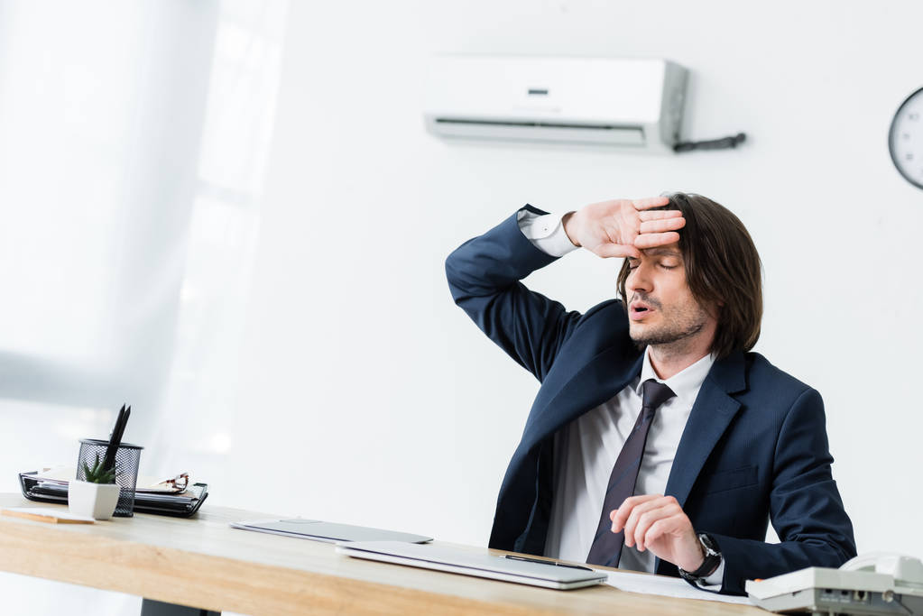 ιδρωμένος επιχειρηματίας κάθεται στο γραφείο, κρατώντας το χέρι κοντά στο πρόσωπο και υποφέρει από τη θερμότητα - Φωτογραφία, εικόνα