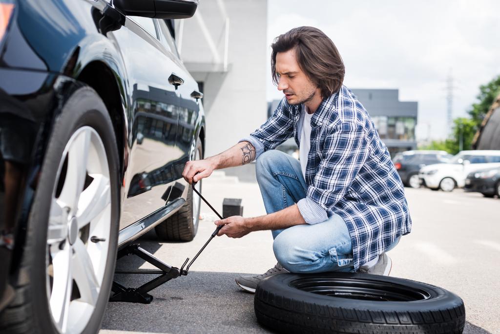 man in casual wear using jack tool and changing broken wheel on auto, car insurance concept - Photo, Image