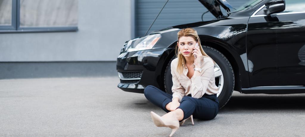 panoramische foto van de overstuur zakenvrouw praten op smartphone en zittend in de buurt van Broken auto, autoverzekering concept - Foto, afbeelding