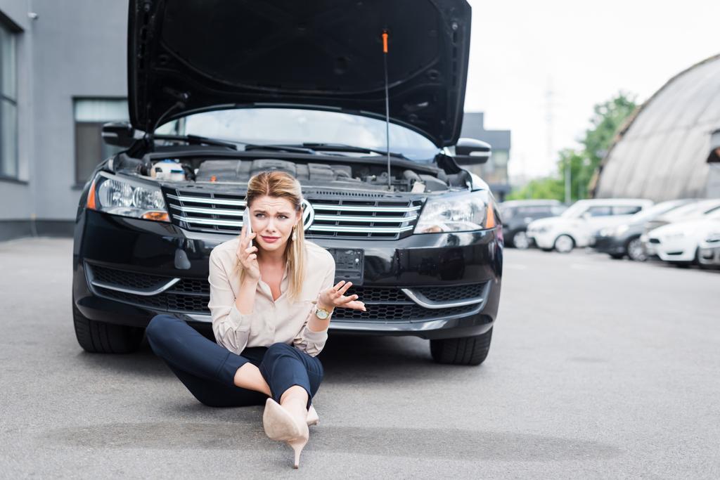 upset businesswoman looking away, talking on smartphone and sitting near broken auto, car insurance concept - Photo, Image