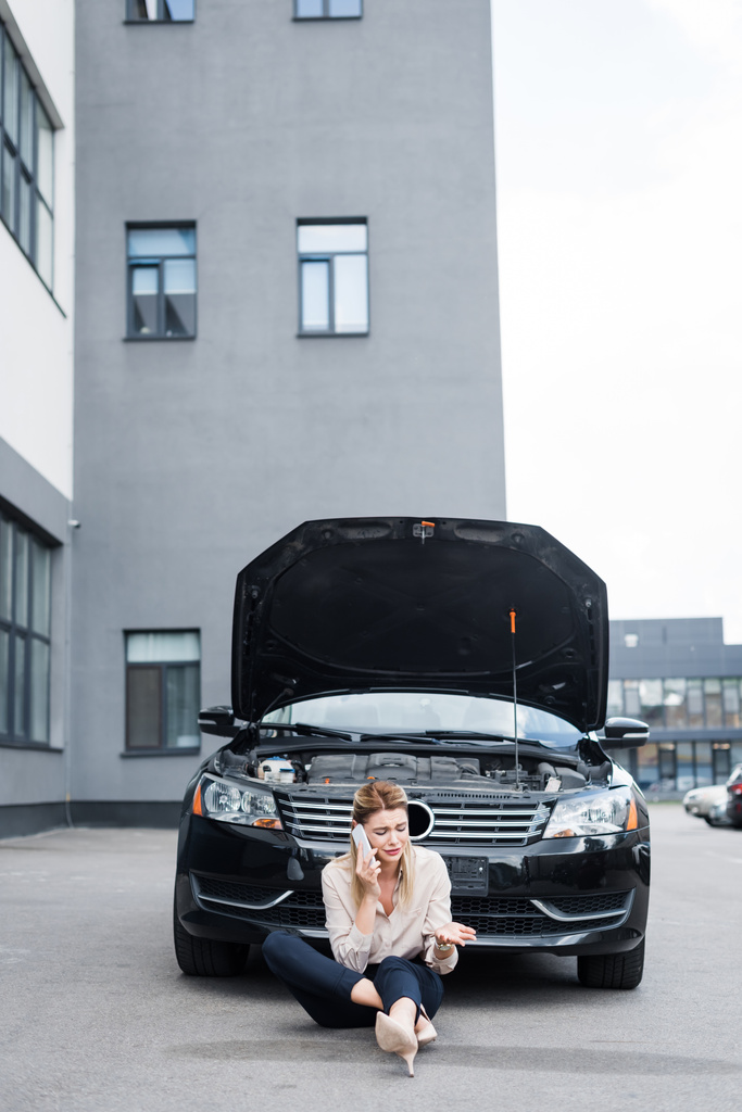 Weinende Geschäftsfrau, die auf dem Smartphone spricht und in der Nähe eines kaputten Autos sitzt, Kfz-Versicherungskonzept - Foto, Bild