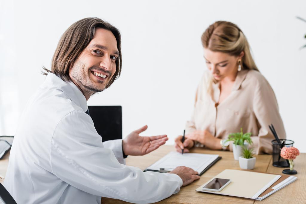 selectieve focus van gelukkige dokter die naar de camera kijkt terwijl het document voor patiënt ondertekening - Foto, afbeelding