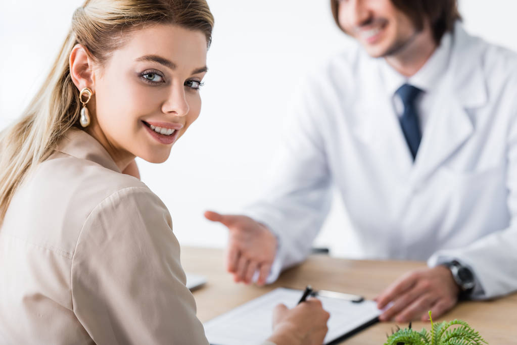 selective focus of patient sitting with doctor behind wooden table, signing document and looking at camera - Photo, Image