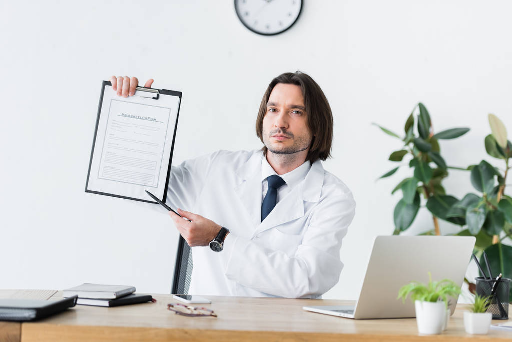 lekarz w białym płaszczu siedzi za drewnianym stołem, patrząc na kamerę i wskazując na pióro w dokumencie medycznym  - Zdjęcie, obraz