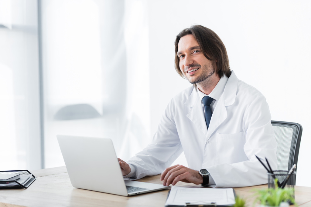 glücklicher Arzt im weißen Kittel, der während der Arbeit mit dem Laptop in die Kamera blickt - Foto, Bild