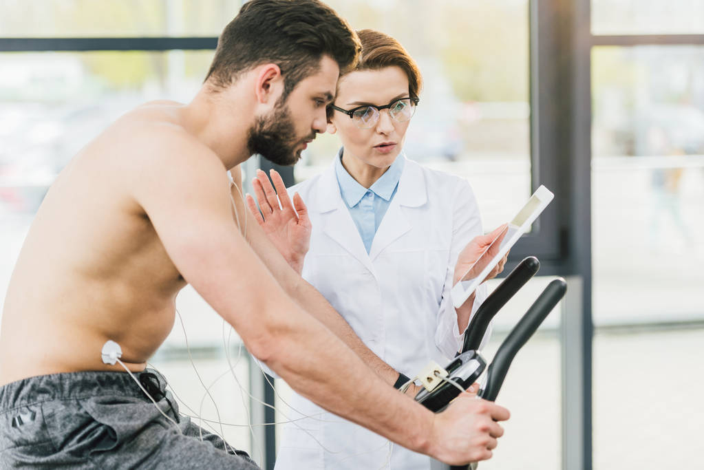 doctor with digital tablet gesturing near shirtless sportsman during endurance test - Photo, Image