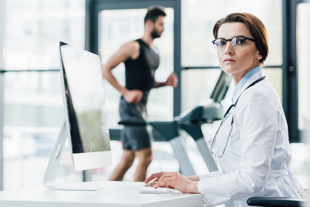 γιατρός σε γυαλιά χρησιμοποιώντας τον υπολογιστή, ενώ αθλητής τρέχει σε διάδρομο κατά τη διάρκεια της δοκιμής αντοχής στο γυμναστήριο - Φωτογραφία, εικόνα