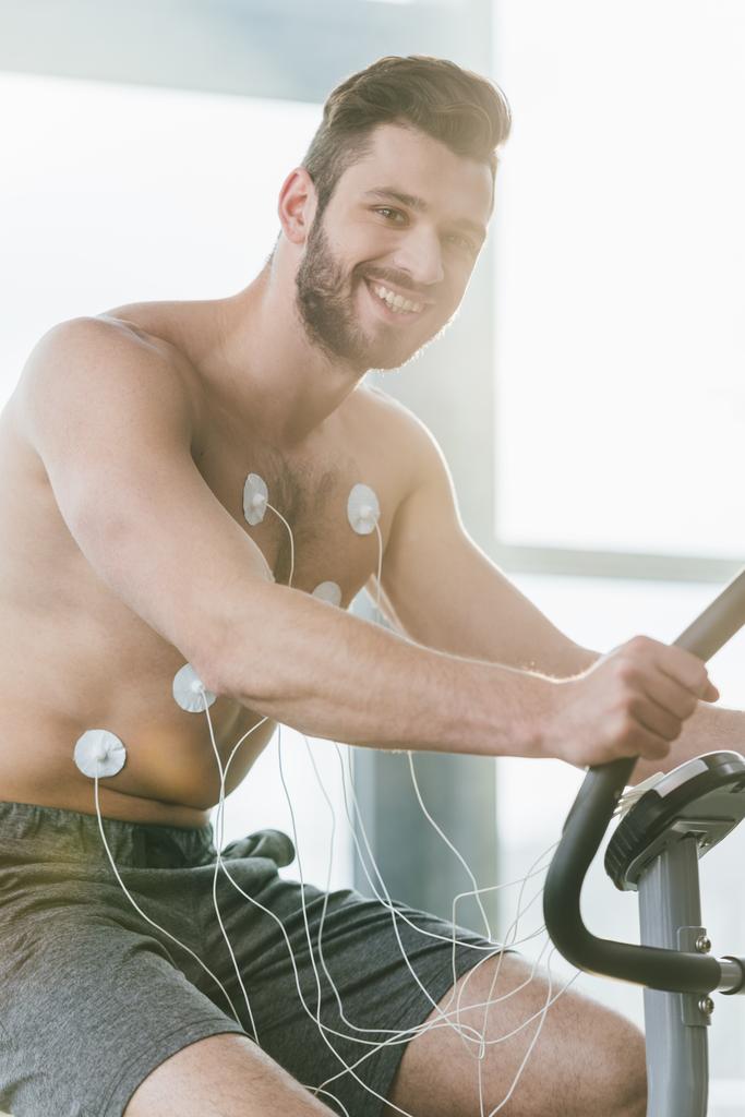 ευτυχισμένος όμορφος αθλητής με ηλεκτρόδια σε ελλειπτικό κατά τη διάρκεια της δοκιμής αντοχής στο γυμναστήριο - Φωτογραφία, εικόνα