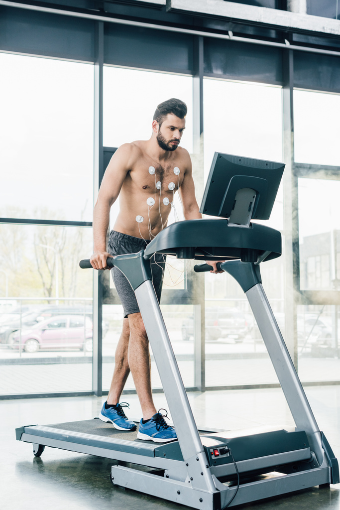 handsome muscular sportsman with electrodes running on treadmill during endurance test in gym - Photo, Image