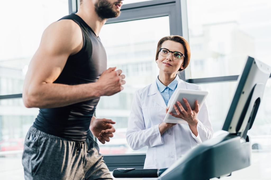 Sportler läuft beim Härtetest im Fitnessstudio mit digitalem Tablet auf Laufband neben Arzt - Foto, Bild