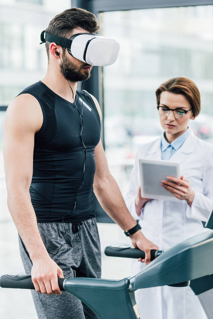спортсмен в гарнітурі vr працює на біговій доріжці біля лікаря під час тесту на витривалість в спортзалі
 - Фото, зображення