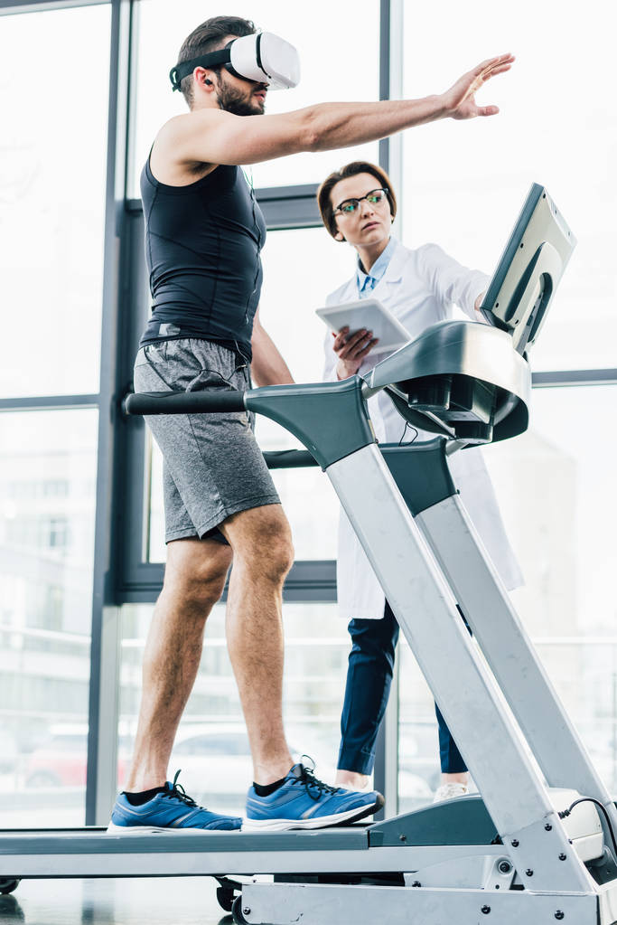 спортсмен в гарнітурі vr жестикулюючи на біговій доріжці біля лікаря під час тесту на витривалість в спортзалі
 - Фото, зображення