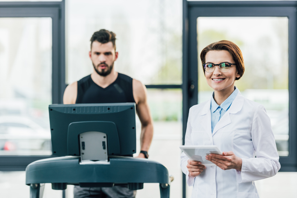 sportovec na běžícím trenažéru blízko usmívajícího se lékaře při zkoušce životnosti v tělocvičně - Fotografie, Obrázek