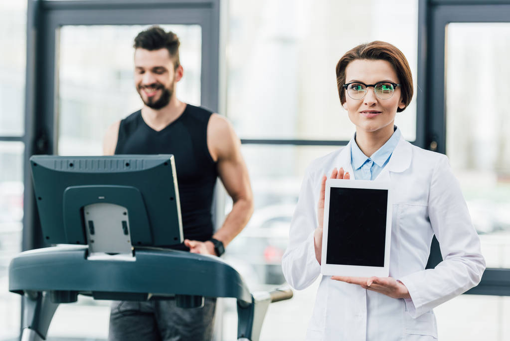 spor salonunda dayanıklılık testi sırasında Dijital Tablet gösteren doktor yakınında koşu bandı üzerinde çalışan sporcu - Fotoğraf, Görsel