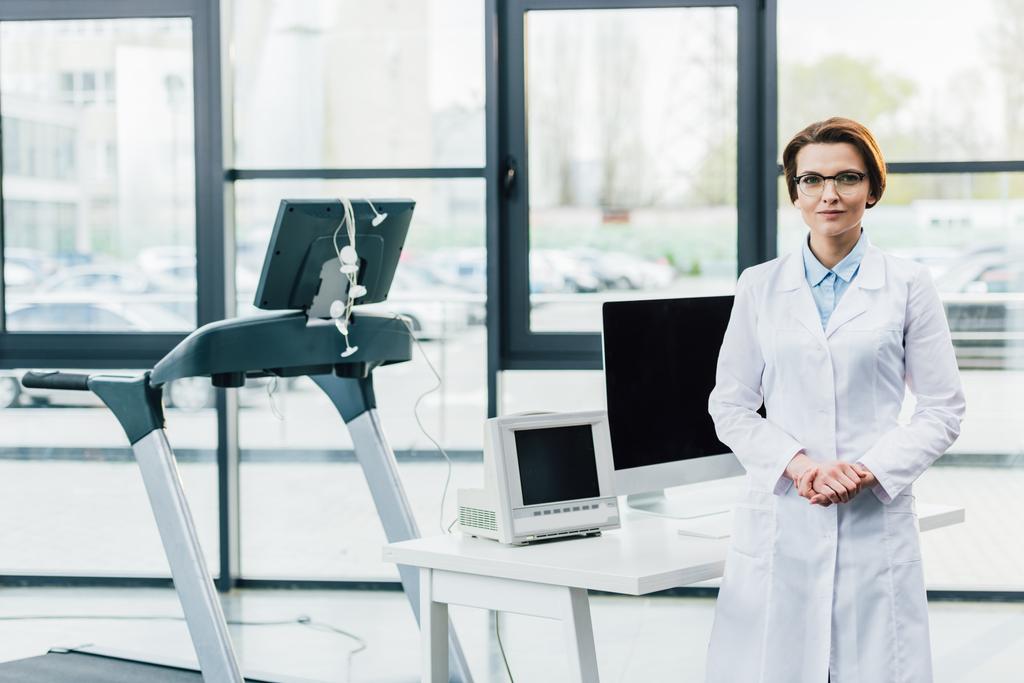 médecin en manteau blanc près du tapis roulant et bureau d'ordinateur à la salle de gym
 - Photo, image