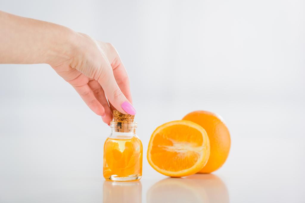 Teilansicht einer Frau, die eine Glasflasche mit ätherischem Orangenöl in der Nähe frischer Orangen auf weißem Hintergrund verkorkt - Foto, Bild