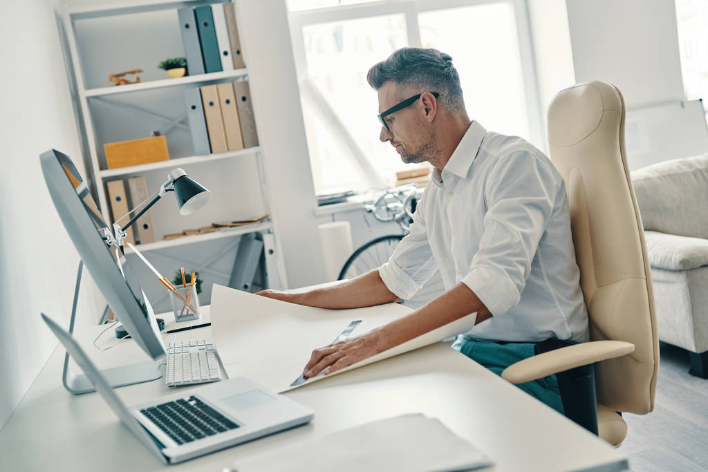 Успешный архитектор, работающий за офисным столом с чертежем, ноутбуком и компьютером
  - Фото, изображение