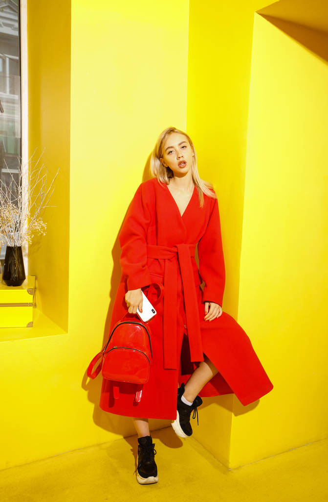 Şık kırmızı ceket giymiş güzel genç kız Blogger göstermek odasında sarı duvarlar arka planda pozlar - Fotoğraf, Görsel