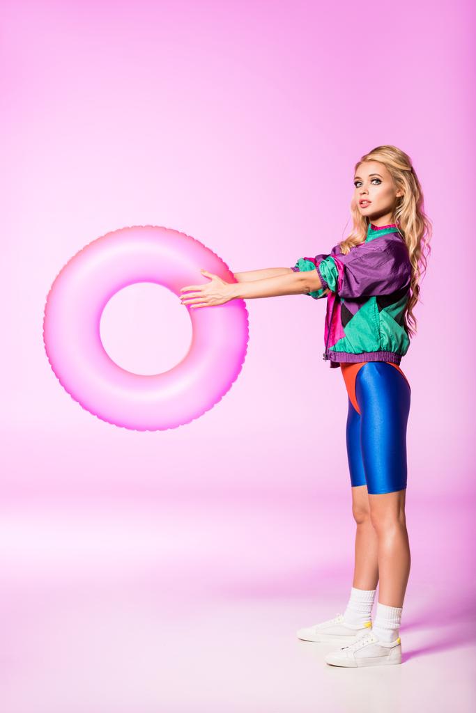 atractiva chica con anillo de natación inflable mirando a la cámara en rosa, concepto de muñeca
 - Foto, Imagen