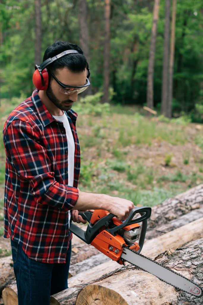 attentive lumberer in checkered shirt cutting wood with chainsaw in forest - Photo, Image