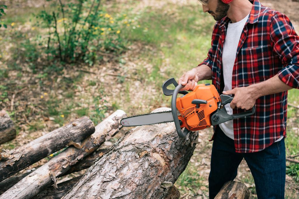 houthakker in geruite overhemd snijden ronde Timbers met kettingzaag in bos - Foto, afbeelding