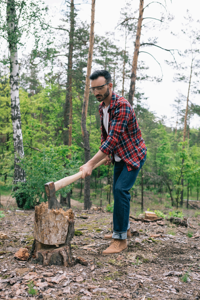 handsome lumberjack in plaid shirt and denim jeans cutting wood with ax in forest - Photo, Image
