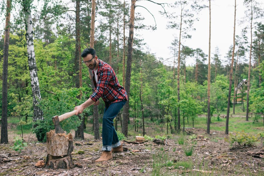 bebaarde lumberman in geruite shirt en denim jeans snijden hout met AX in bos - Foto, afbeelding