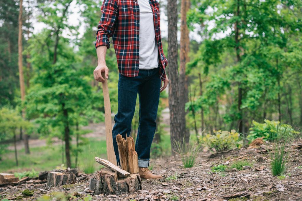 částečný pohled na dřevorubec v Kostkové košili a džínsy stojící s sekyrou v lese - Fotografie, Obrázek