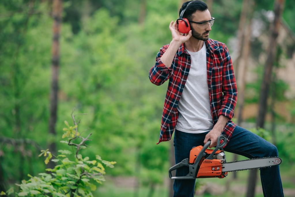 ξυλοκόπος σε καρό πουκάμισο στέκεται με αλυσοπρίονο στο δάσος και αγγίζοντας τα ακουστικά ακύρωσης θορύβου - Φωτογραφία, εικόνα